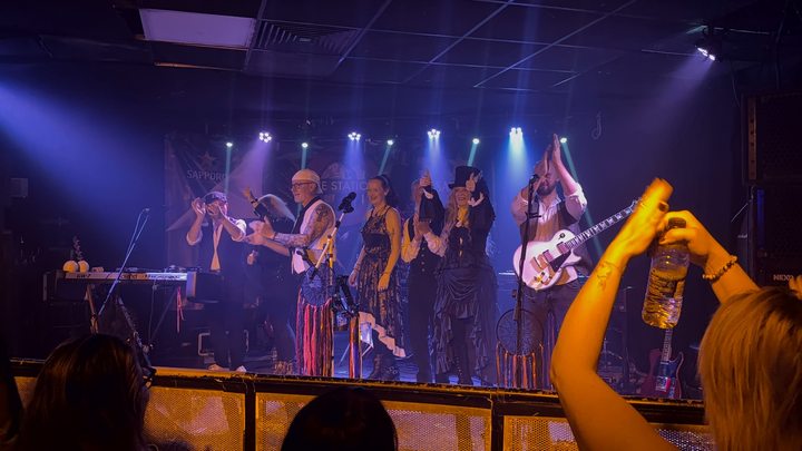 Fleetwood Mad performing beneath bright stage lights as someone in the crowd raises their hands
