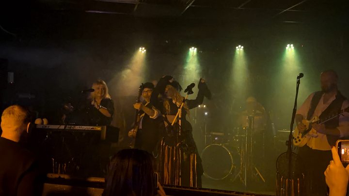 Fleetwood Mad singer raising one hand at the microphone with a guitarist beside her under warm stage lights