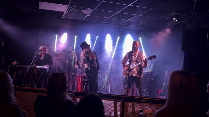 Fleetwood Mad performing with singers and a guitarist beneath bright white stage beams